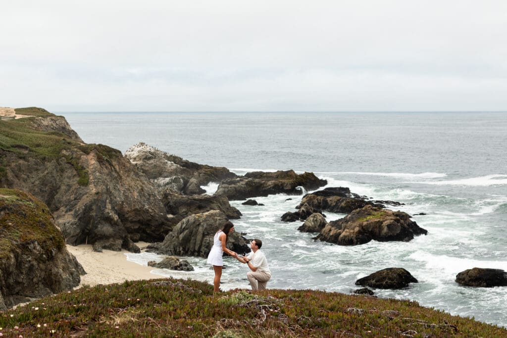 A man kneels to propose to his girlfriend at Bodega Head with a view of the ocean.