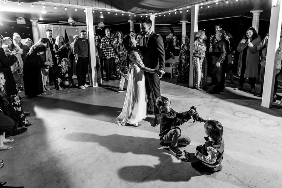 Black and white photo of bride and groom dancing at wedding reception with kids dancing on the floor