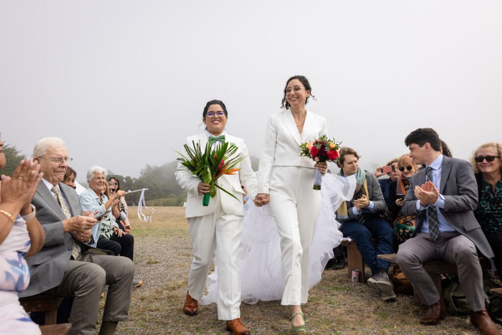 Brides walking down the aisle with bouquets and wedding guests in Holly’s Ocean Meadow in Fort Bragg, CA